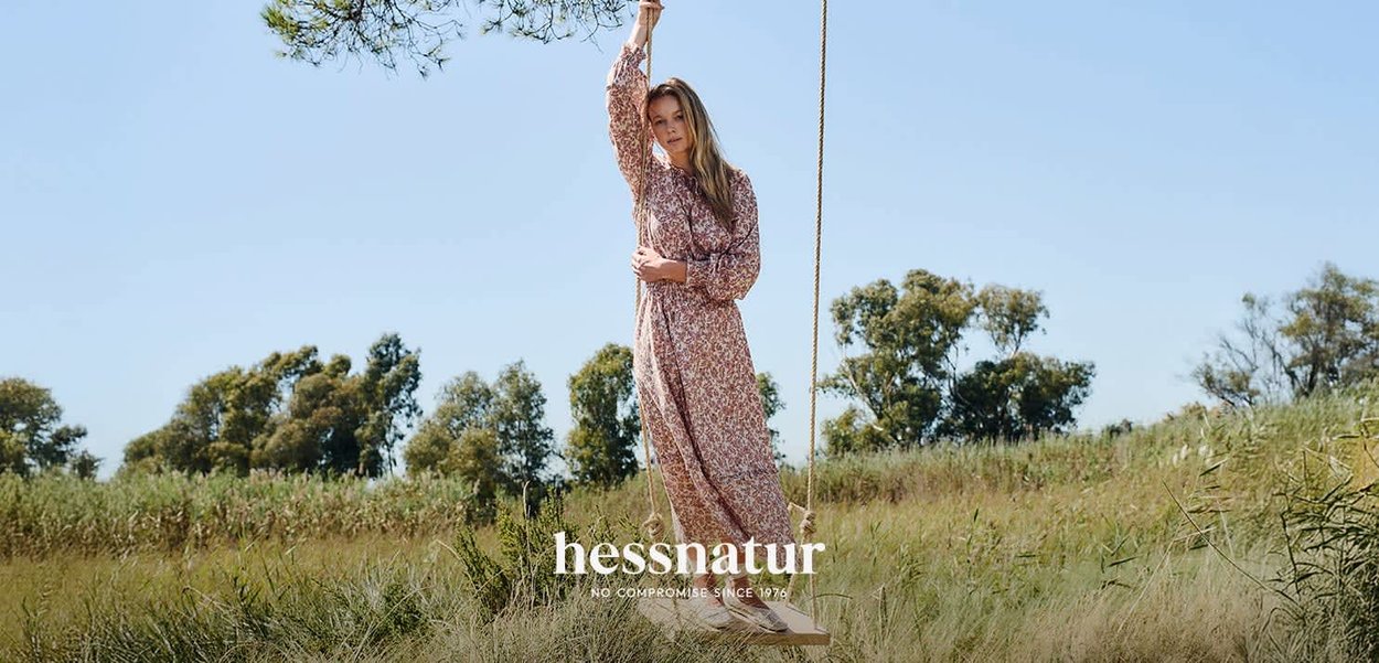 Une femme en robe longue à fleurs est debout sur une balançoire en bois suspendue à un arbre dans un champ herbeux avec des arbres en arrière-plan.
