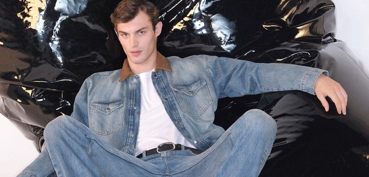 Giacca di jeans con colletto marrone, t-shirt bianca e jeans azzurri chiari. Seduto su una superficie nera lucida, postura rilassata, sfondo neutro.