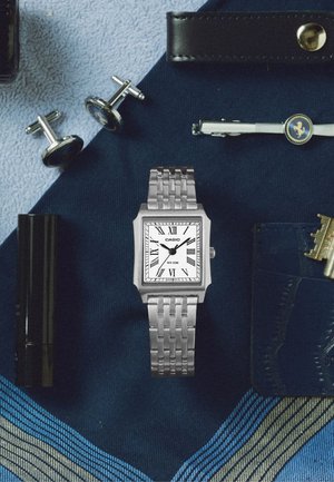 Zilveren Casio polshorloge met witte wijzerplaat en Romeinse cijfers, omringd door manchetknopen, leren portemonnee, dasspeld, sleutels en gestreepte stropdas op stof.
