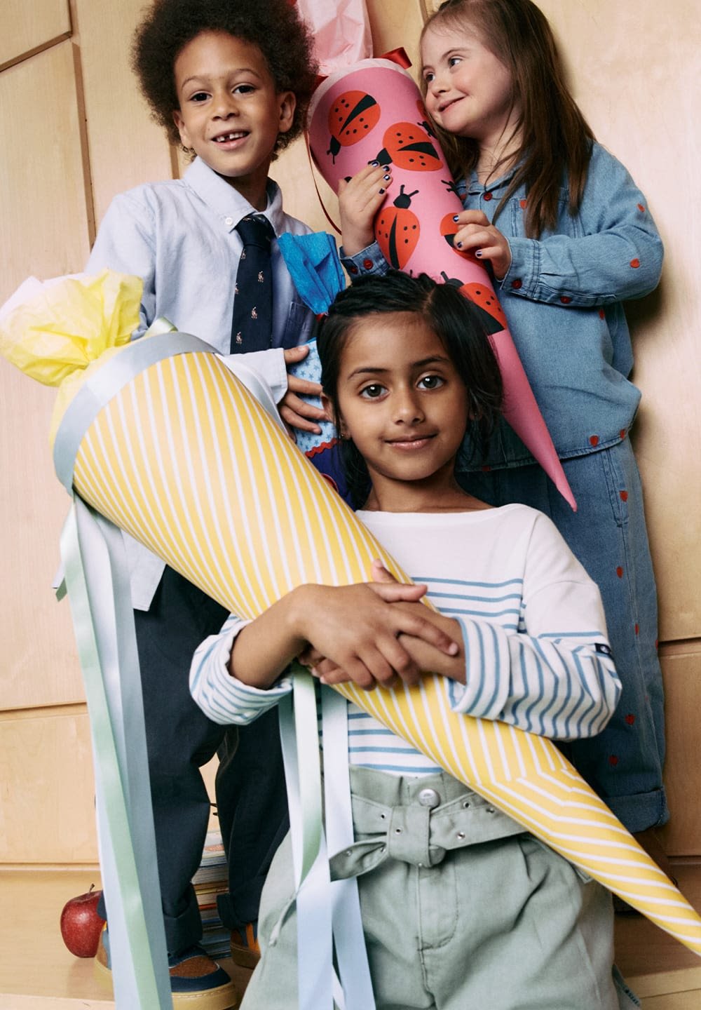 Three children hold colorful cone-shaped gift bags. One bag is yellow with white stripes, another is pink with ladybug patterns, and a blue bag is visible.