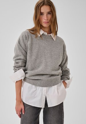 Grauer Strickpullover mit rundem Ausschnitt und geripptem Saum, getragen über einem weißen Hemd mit Kragen. Vor einem schlichten, hellen Hintergrund.