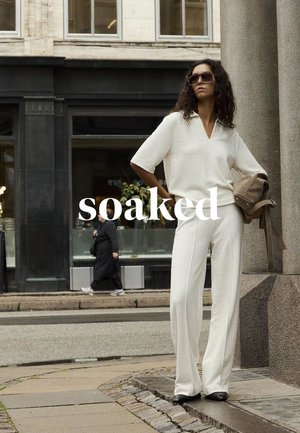 Kvinde iført hvidt outfit og solbriller læner sig mod en stensøjle og holder en beige taske på en byfortov med bygninger i baggrunden.