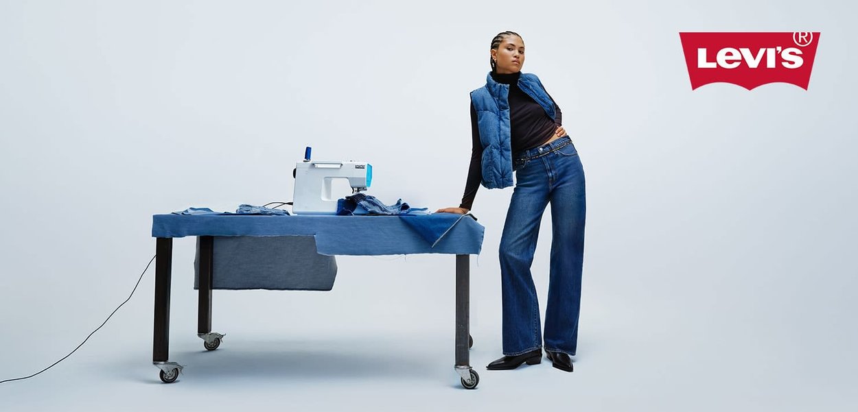 Blå denim bell-bottom bukser kombinert med en svart turtleneck og en blå hettevest. En symaskin og stykker med stoff vist på et bord.