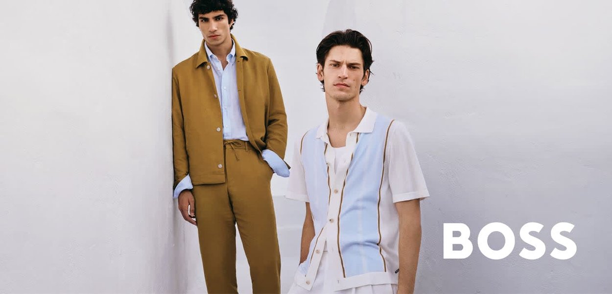 Due giovani uomini indossano abiti alla moda, uno in completo beige con camicia azzurra, l'altro con camicia e pantaloni casual bianchi e azzurri.