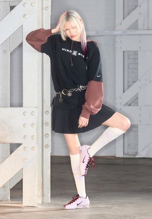 Jong persoon met blond en donker haar in een zwarte Nike-hoodie, geplooide rok, witte kniekousen en lila sneakers, poserend binnen bij metalen balken.