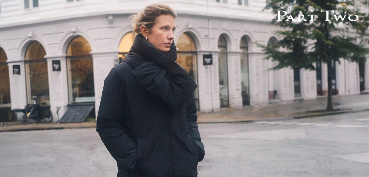 Schwarze Steppjacke mit großem Kragen, die eine strukturierte Oberfläche und zwei Seitentaschen aufweist. Im Hintergrund ist eine Stadtstraße mit Bögen zu sehen.