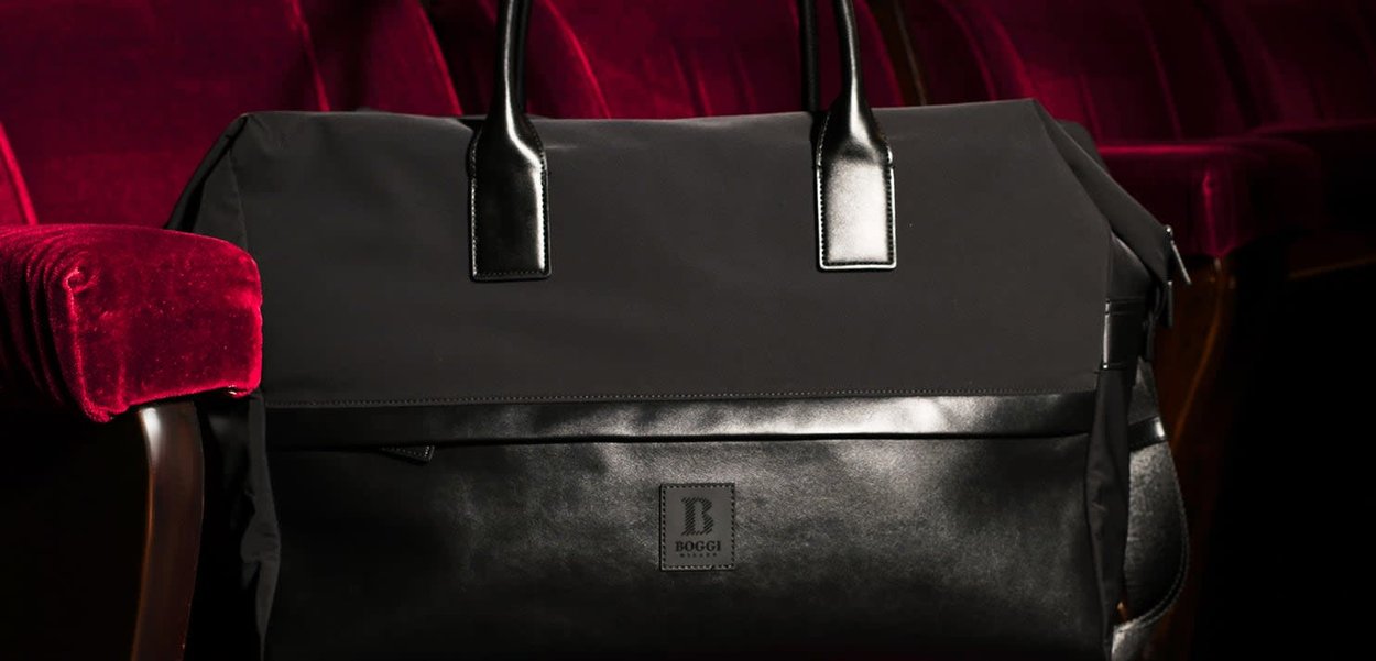 Sac de sport noir en tissu mat, avec des accents en cuir brillant et des poignées robustes. Comprend un patch logo et une poche zippée.