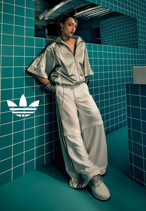 Zilveren oversized jas met de tekst "TEAMAGE", gecombineerd met witte wijde broek met zwarte zijstrepen en grijze sportschoenen.