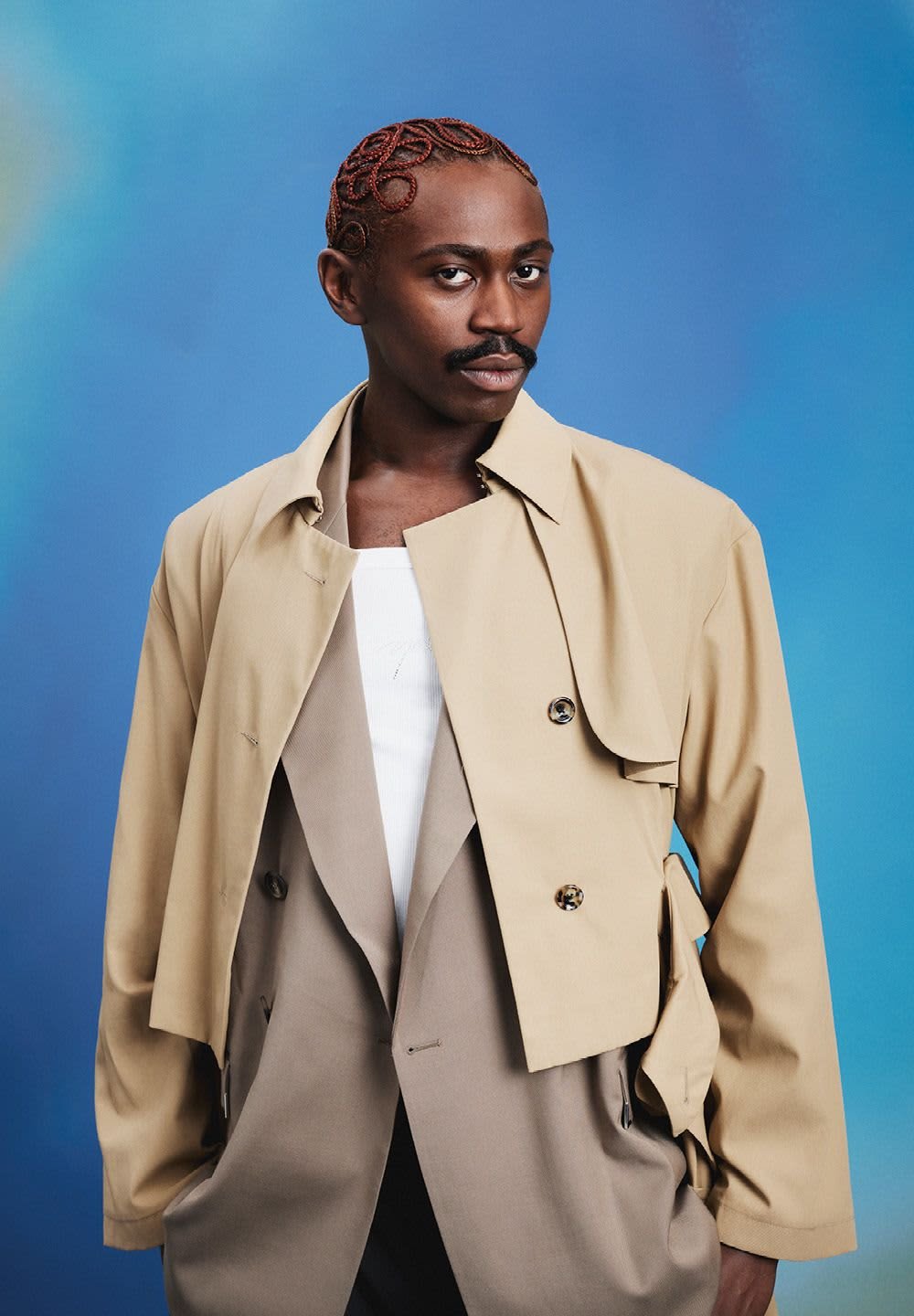 Beige trenchcoat med överdimensionerad krage och framknappar, lager på ett ljusgrått plagg, satt mot en blå bakgrund.