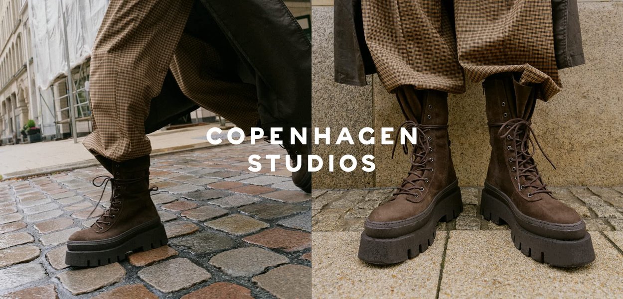 Bruine suède chunky platform veterschoenen gedragen met bruine geplooide brede broeken tegen een achtergrond van een stenen muur en asfalt.