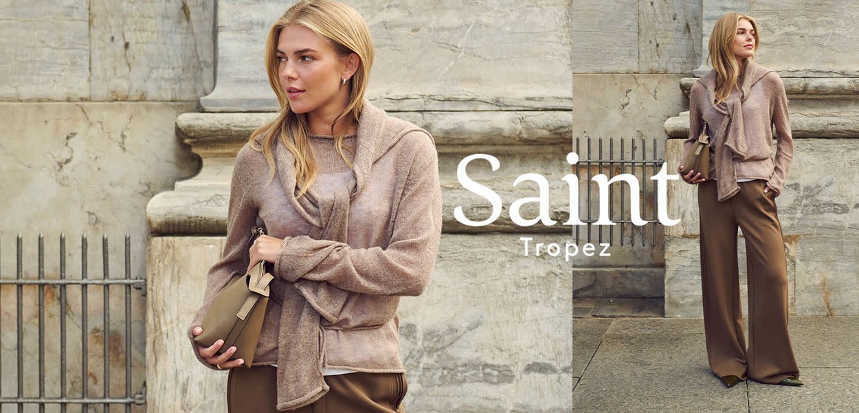 Vrouw die een taupekleurige trui en brede bruine broek draagt, houdt een beige tas vast, staand tegen een stenen muur met nabij de tekst Saint Tropez.