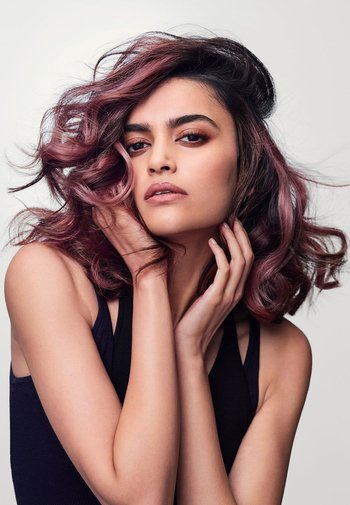 Model with wavy, shoulder-length hair featuring a gradient from dark to light pink. She's wearing a black sleeveless top, posing with hands near her face.
