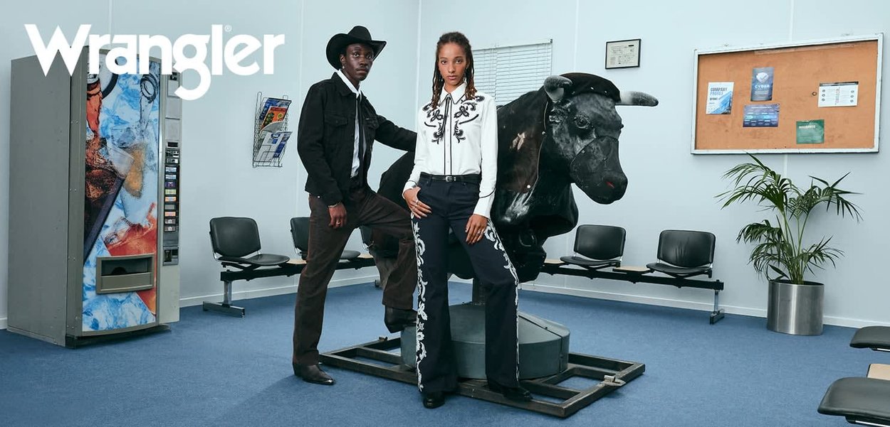 Zwei Models posieren mit einem mechanischen Bullen. Der eine trägt einen schwarzen Cowboyhut, eine Jacke und dunkle Hosen; der andere trägt ein weißes Hemd mit schwarzer Stickerei und Jeans mit ähnlichen Akzenten.