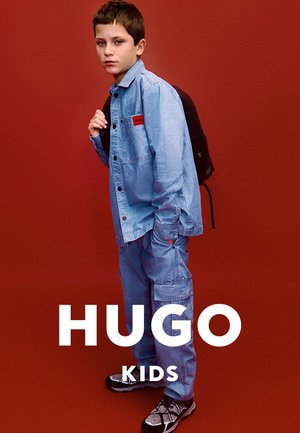 Veste en jean et pantalon bleu clair, avec un patch de logo de marque rouge, associés à des baskets noires et grises. Le modèle tient un sac à dos noir.