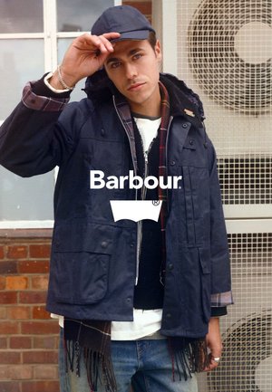 Marineblauer wasserdichter Mantel mit mehreren Taschen, einem karrierten Futter und einer Kapuze. Getragen über einem schwarzen Pullover und einem weißen Hemd. Begleitet von einer marineblauen Kappe.