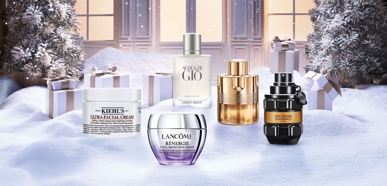 Five cosmetic products on snow-covered ground: Kiehl's Ultra Facial Cream (white jar), Acqua di Gio (clear bottle), Lancome Renergie (silver and purple jar), a gold bottle, and Spicebomb (black bottle with a tag).