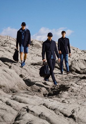 Drei Männer gehen über felsiges Gelände und tragen dunkle Jacken sowie blaue Hosen oder Shorts, zwei von ihnen tragen schwarze Taschen, unter einem klaren blauen Himmel.