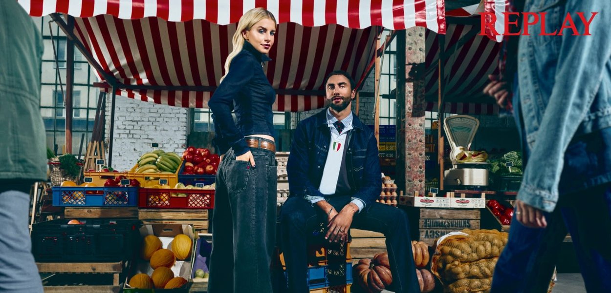 En blond kvinde står med hænderne i lommerne, mens en mand sidder iført denim-outfits i et indendørs marked fyldt med frugter og grøntsager.