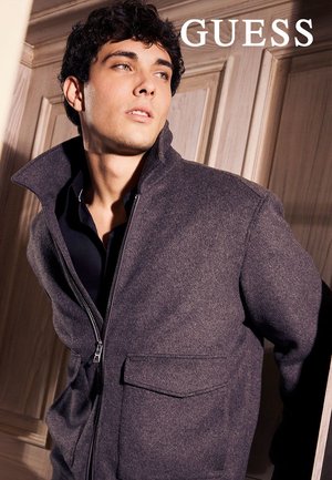 Giacca grigia con cerniera e collo alto, tessuto testurizzato, due tasche frontali e design elegante. Logo del marchio "GUESS" visibile in alto.