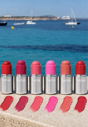 Sei rossetti TIRTIR in tonalità rosse, rosa e nude con campioni abbinati, posizionati su un bordo di pietra accanto a un mare blu calmo con barche sullo sfondo.