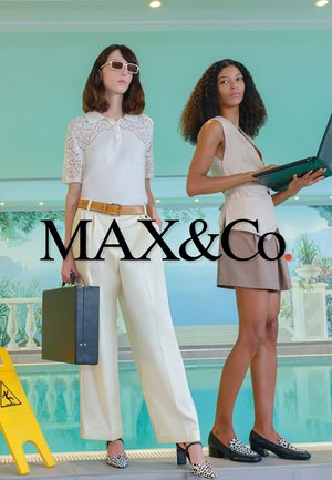 Zwei Frauen neben einem Hallenbad, eine trägt weiße Hosen und Sonnenbrillen und hält eine Aktentasche, die andere trägt ein beiges Outfit und hält einen Laptop.