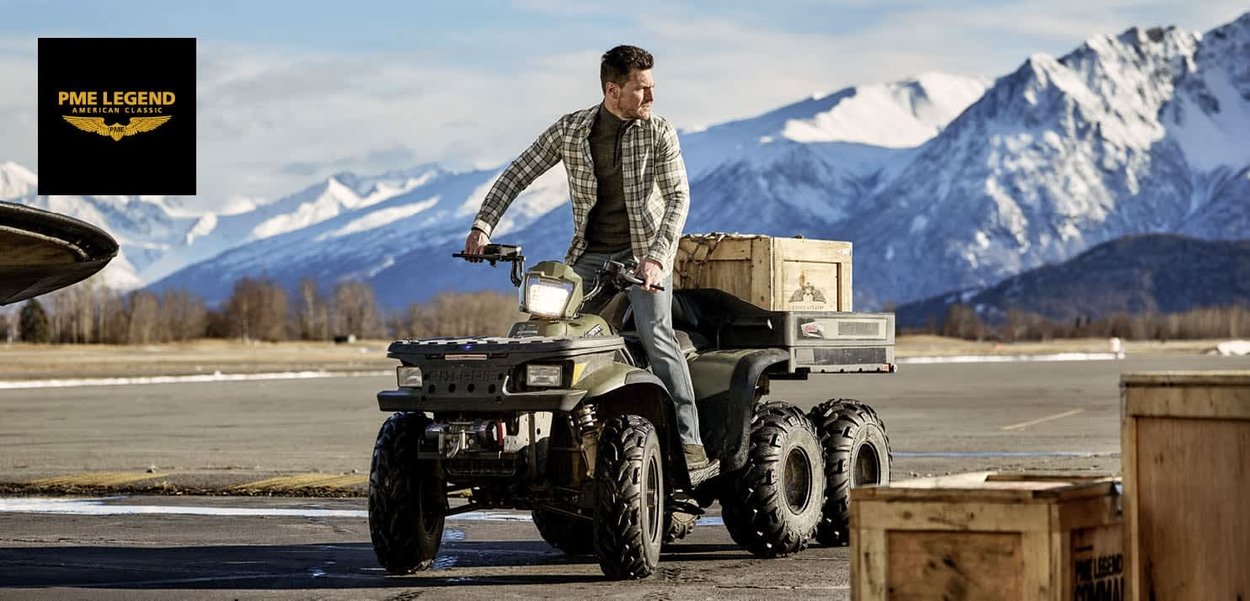 Veicolo tutto terreno di colore verde con grandi pneumatici neri, un portapacchi anteriore e casse di legno nelle vicinanze. Lo sfondo presenta montagne innevate.