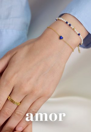 Goldene Kettenarmband mit einem blauen Herzanhänger, begleitet von einemPerlenarmband mit weißen Perlen und blauen Akzenten sowie einem goldenen Glieder-Ring.