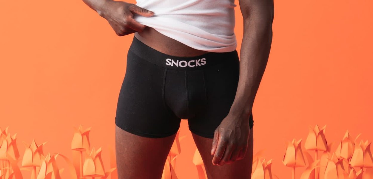 Mand løfter hvid skjorte for at afsløre sorte Snocks boxershorts mod en orange baggrund med abstrakte blomsterformer.
