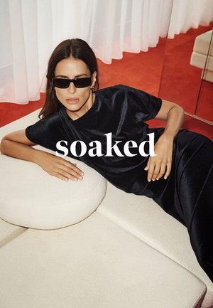 Black satin outfit featuring a relaxed fit. Model wears dark sunglasses, resting on a cream circular cushion against a red carpet backdrop.