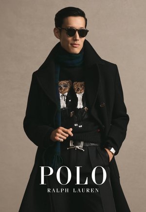 Mann trägt schwarzen Mantel, Bärenpullover, grünen Schal, Sonnenbrille und Uhr, eine Hand in der Tasche und die andere hält das Revers des Mantels.