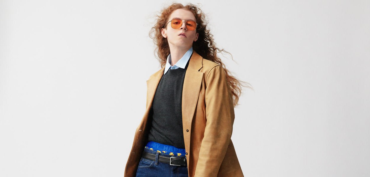 Brown leather coat, gray sweater, blue collared shirt, blue belt with text, dark jeans, and orange sunglasses. Wavy hair, neutral background.