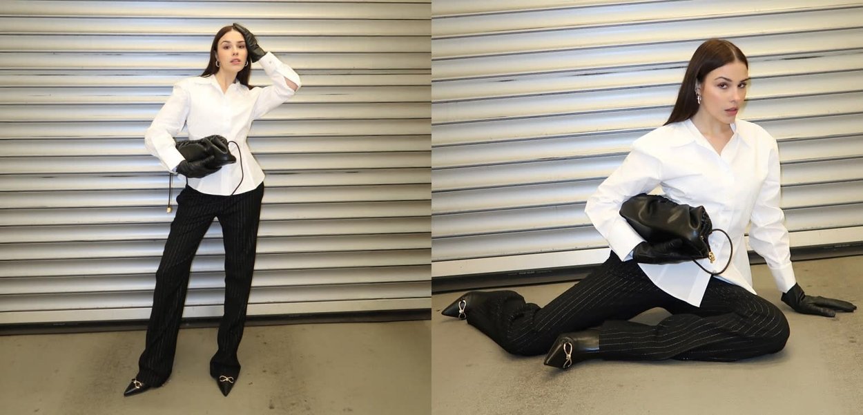 Camisa blanca abotonada, pantalones negros con rayas finas, guantes de cuero y un bolso negro con un anillo dorado. Corte ajustado y aspecto pulido, con detalles mínimos.