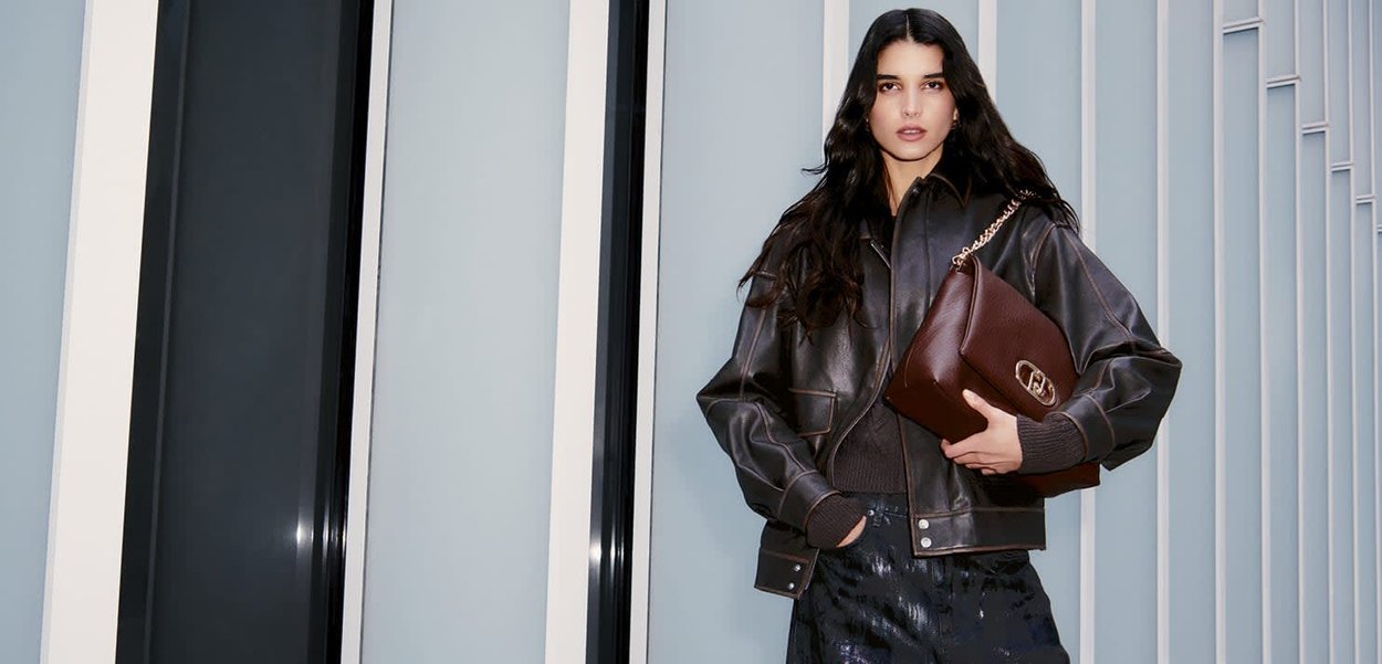 Giacca in pelle marrone con design oversize, polsini a coste e dettagli di cucitura; una borsa marrone abbinata con un accento dorato.