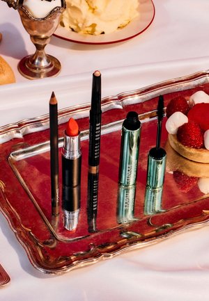 Quatre produits cosmétiques sur un plateau miroir argenté : un crayon marron, un rouge à lèvres orange, un eye-liner noir et un tube de mascara vert, présentés de manière ordonnée.