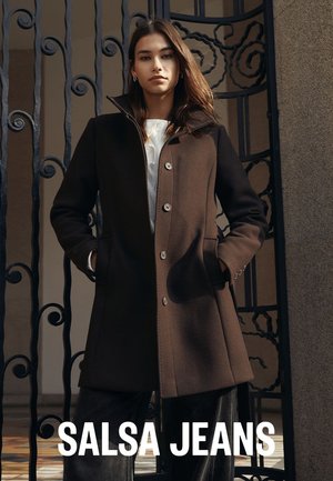 Brauner Wollmantel mit hohem Kragen und silbernen Knöpfen, mit zwei Seitentaschen und tailliertem Schnitt, getragen über einem weißen Oberteil.