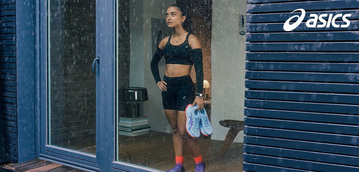 Black athletic crop top and shorts, orange and purple socks, holding light blue running shoes, standing behind a rain-streaked glass door.