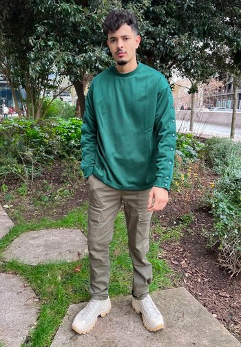 Grön långärmad sweatshirt med knappdetaljer, tillsammans med kaki cargobyxor och klumpiga beiga sneakers, stående på en stenbelagd väg i grönskande omgivningar.