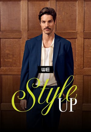 Man with mustache wearing navy blue suit and white shirt, standing against wooden paneled wall with "WE Style UP" text overlay.