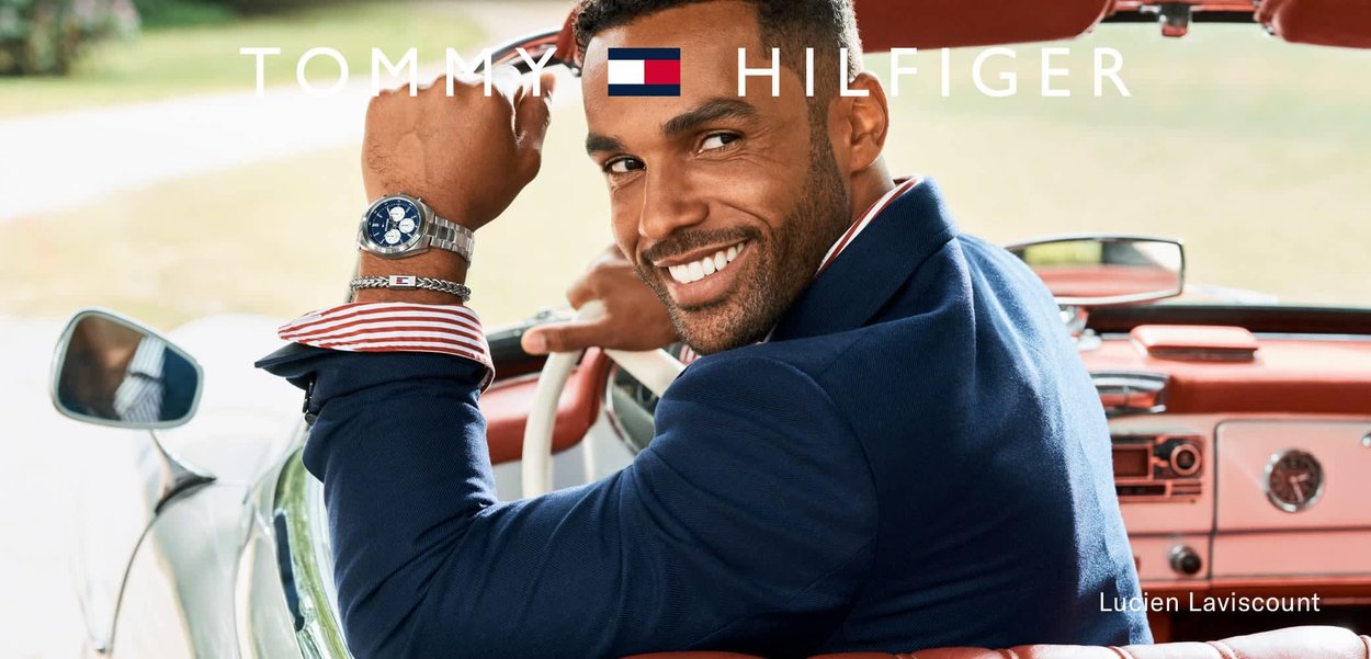 Homme souriant en blazer bleu marine et chemise rayée conduisant une voiture vintage, portant une montre argentée et un bracelet avec le logo Tommy Hilfiger.