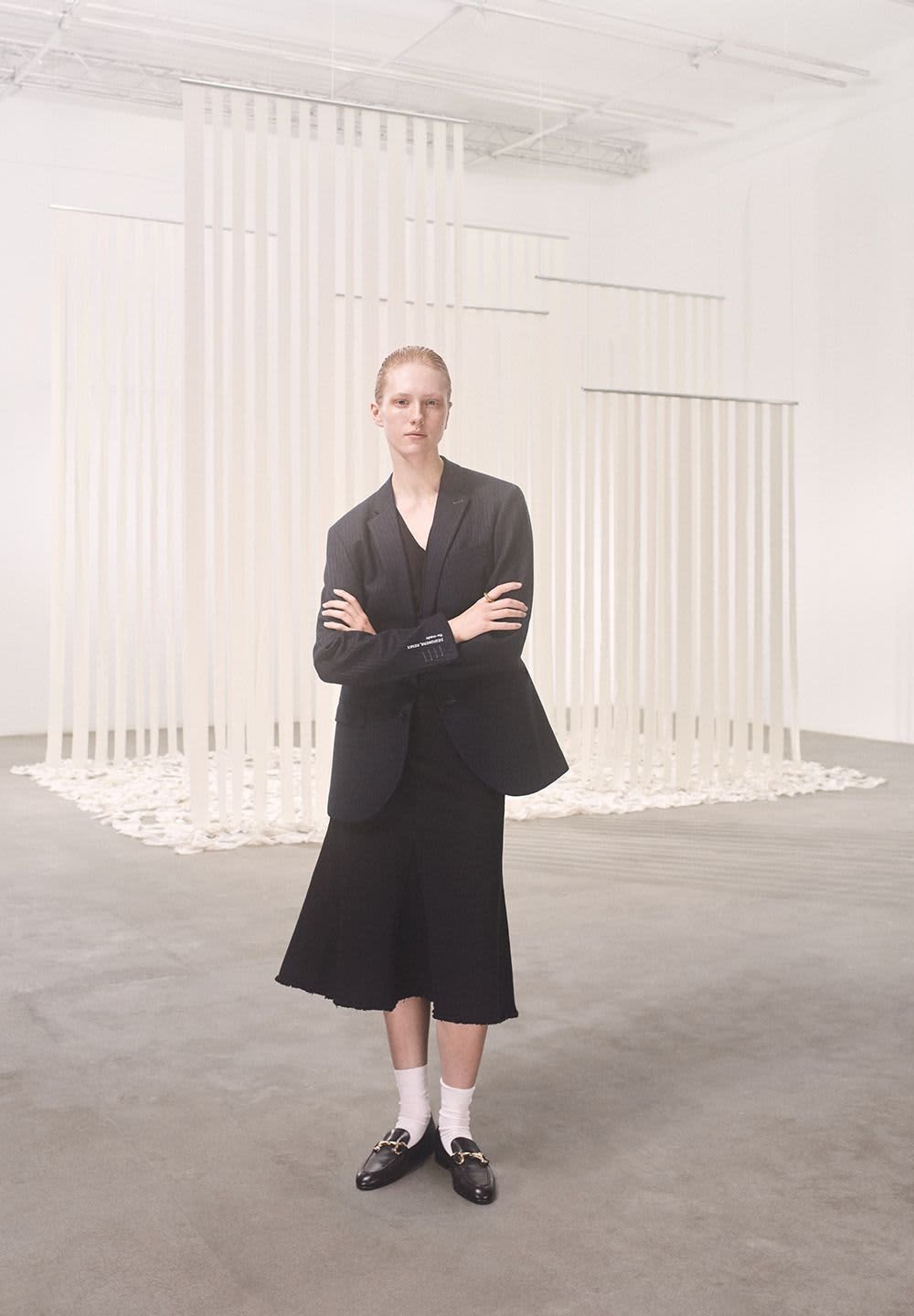Black tailored blazer paired with a black skirt, white socks, and black loafers; stands in front of vertical white fabric strips.