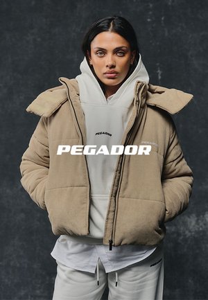 Frau trägt eine beige gesteppte Jacke über einem weißen Hoodie und weißen Hosen, auf denen der Markenname "PEGADOR" sichtbar ist, vor dunklem Hintergrund.