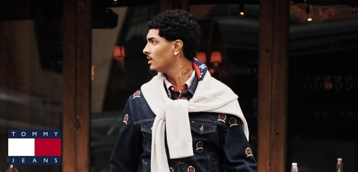 Chaqueta de mezclilla con acentos bordados, combinada con una camisa de cuadros roja y blanca. Suéter blanco dejado caer sobre los hombros. Cabello negro.