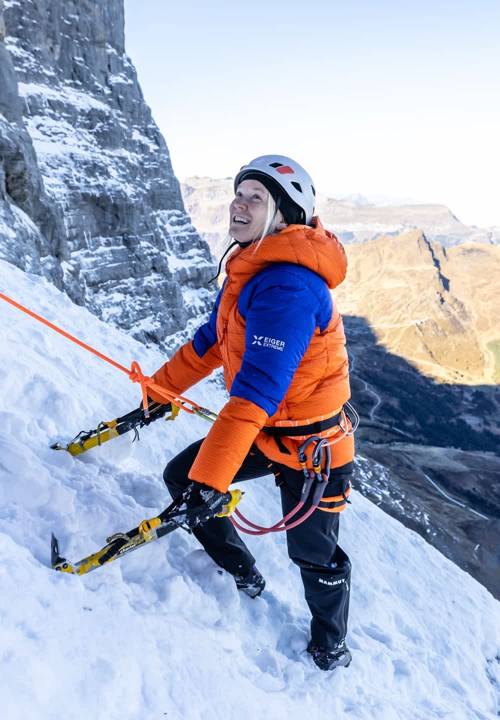Kletterer in orange-blauer Jacke erklimmt schneebedeckte Felswand mit Eisschrauben, gesichert mit Seilen und einem Helm tragend.