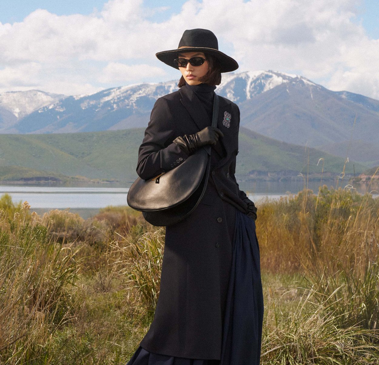 Sac en cuir noir de forme arrondie, associé à un manteau foncé et des gants. Le modèle porte un chapeau noir à large bord et des lunettes de soleil.