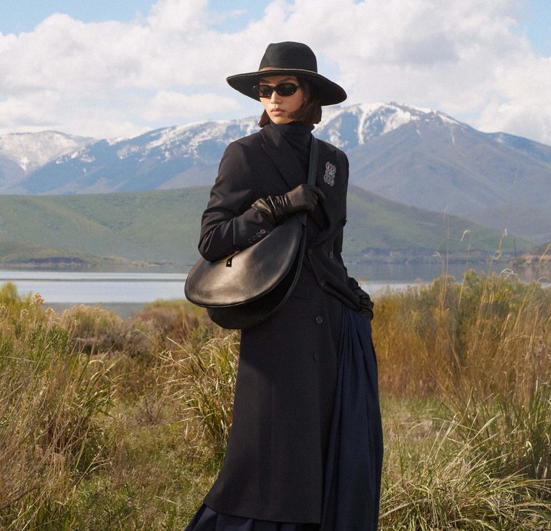 Sac en cuir noir de forme arrondie, associé à un manteau foncé et des gants. Le modèle porte un chapeau noir à large bord et des lunettes de soleil.