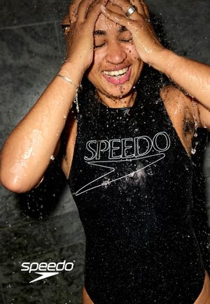 Person in einem schwarzen Speedo-Badeanzug lächelt mit geschlossenen Augen unter fließendem Wasser, Hände am Kopf, Tropfen spritzen herum.