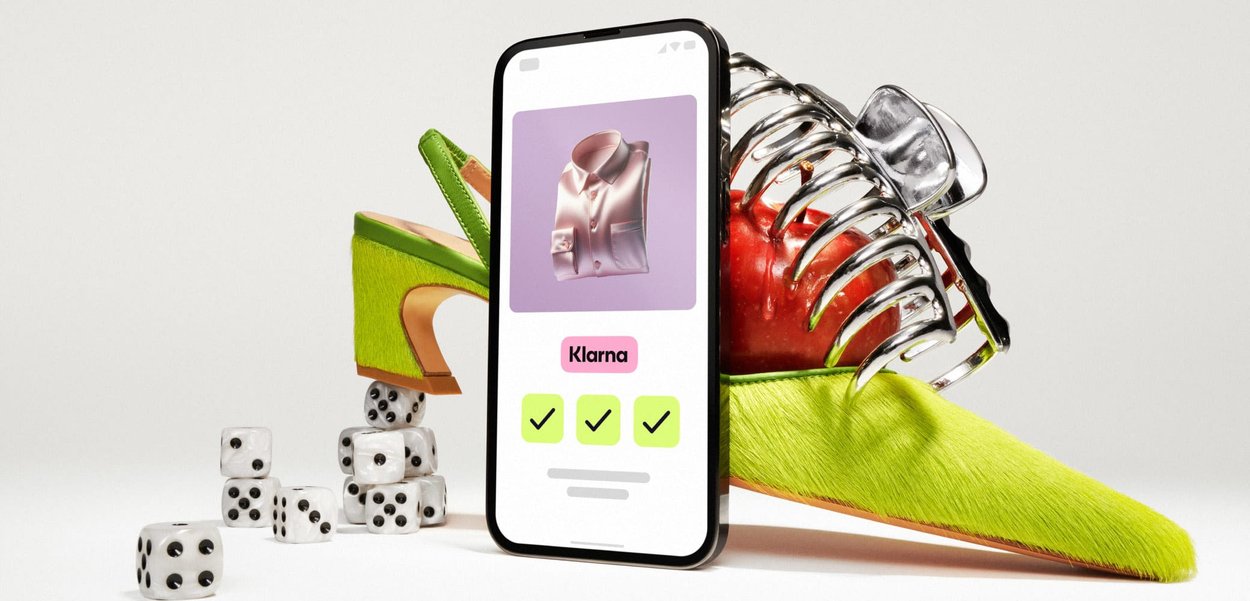 Smartphone met daarop een opgevouwen shirt in de Klarna-app, omgeven door een groene hoge hak op dobbelstenen en een tomaat die vastgehouden wordt door een chroomkleurige klauw.