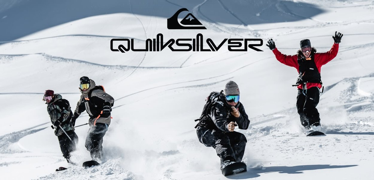 Vier Snowboarder fahren durch frischen Schnee und tragen bunte Ausrüstung. Im Hintergrund sieht man sanfte weiße Hänge unter klarem Himmel. Quiksilver-Logo sichtbar.