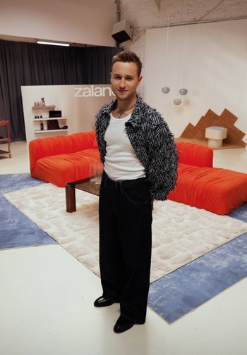 Jeune homme en pantalons larges noirs, débardeur blanc et veste texturée, debout dans une pièce moderne avec des canapés orange et une décoration géométrique.