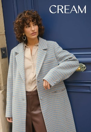 Manteau oversize à carreaux crème et bleu avec un col cranté, une poche avant et des détails de boutons, porté sur un chemisier beige et un pantalon marron.